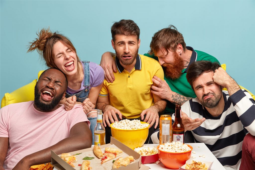 Horizontal shot of funny friends watch humor TV show, express different emotions, enjoy comedy film, eat delicious popcorn and pizza, drink beer, have fun at home, gather together on weekend